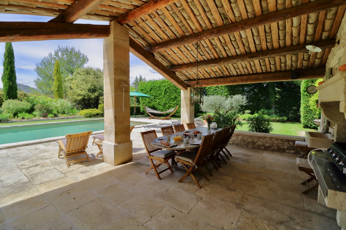 A charming stone farmhouse surrounded by lush greenery under a clear sky, featuring an arched entryway and a covered patio area with outdoor seating.